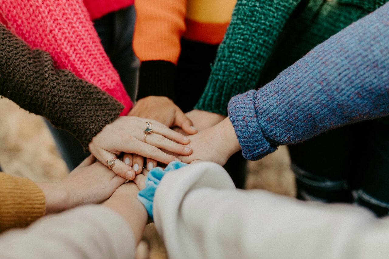 photo of a group of people standing in a ring with their hands pressed together into the middle to signify teamwork and friendship