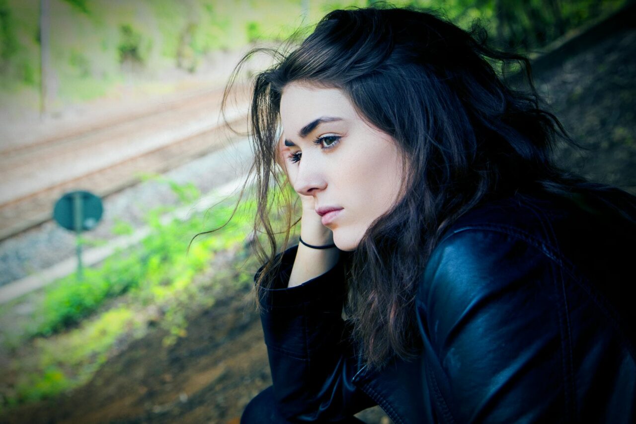 photo of a n attractive young European woman with black straight hair and a black leather jacket waiting for a train